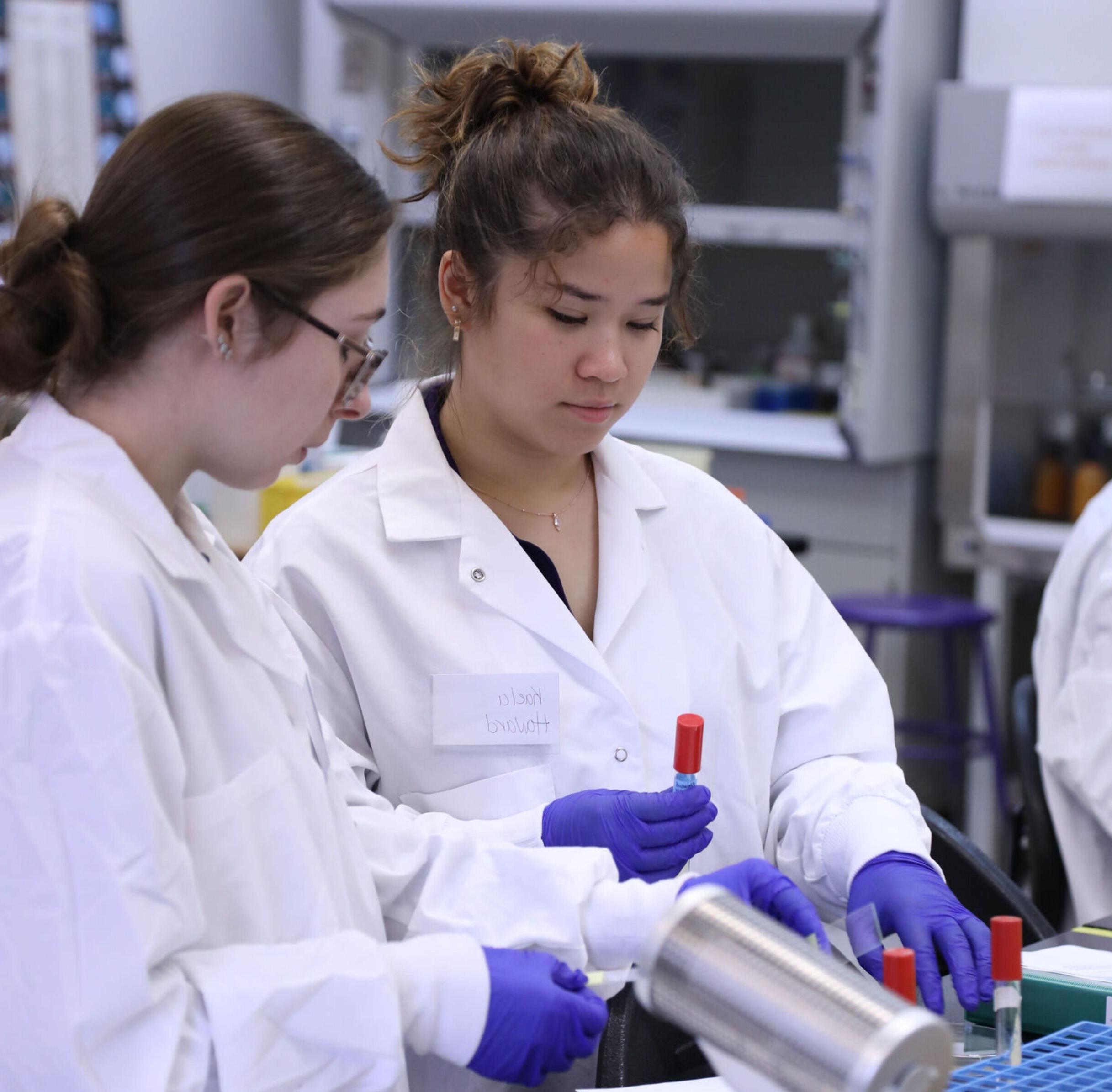 Two female students in lab coats work with vials in a laboratory